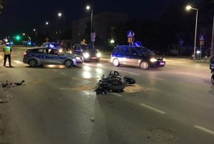 policjant obsługuje zdarzenie z udziałem motocyklisty. W tle radiowóz, motocykl i inne pojazdy