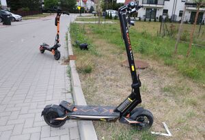 hulajnoga stoi częściowo na chodniku i trawniku, z tyłu druga hulajnoga stoi na chodniku,o obok leży kask